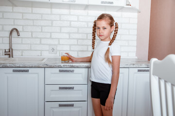 Fototapeta na wymiar Little girl drinks fresh orange juice from a glass cup in the kitchen