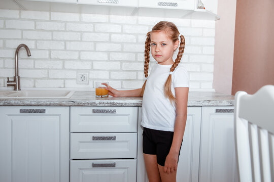 Little girl drinks fresh orange juice from a glass cup in the kitchen