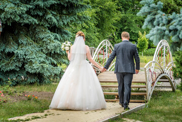 The bride in white wedding dresses and the groom in a tuxedo are walking on the bridge. Happy loving couple on wedding day outdoors