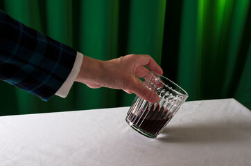 Bartender wearing white blouse and jacket holding glass of red sweet wine, white tablecloth and green curtains