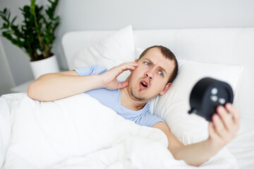 overslept shocked man looking at alarm clock lying in bed at home