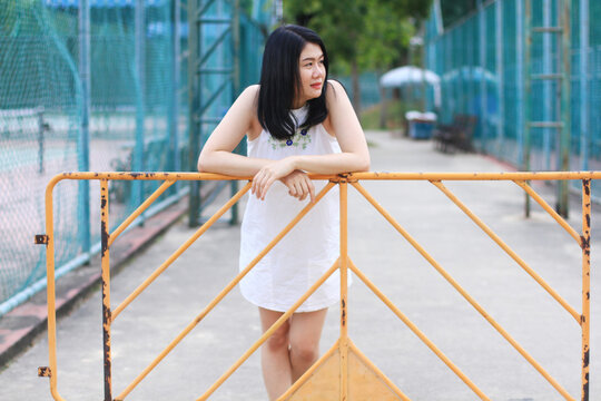 Beautiful asian woman in white dress smiling and stand with yellow steel barrior and blue grating iron fence in natural park. Young Thai girl enjoy on holiday with sunlight in the garden.