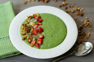 Green smoothie bowl with banana, strawberry, spinach, kale, kiwi, coconut milk garnished with homemade oat, nut and seed granola, coconut flakes, chia seeds, kiwi and strawberry