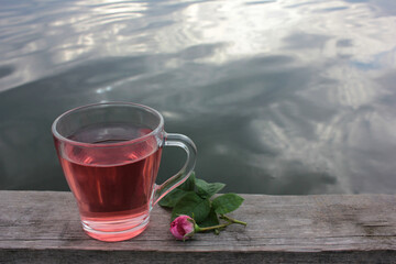 Glass cup of herbal tea with a pink tea rose petals on the wooden pier over the river. Summer tea party outdoors