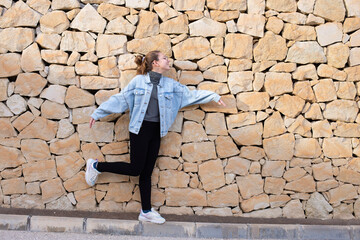Fototapeta na wymiar Young beautiful happy stylish hipster girl in denim jacket and glasses smiling having fun, teenager, stone wall background. Copy space