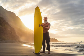 Happy fit senior having fun surfing at sunset time - Sporty bearded man training with surfboard on the beach - Active elderly people lifestyle and extreme sport concept - obrazy, fototapety, plakaty