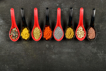 Top view of different kinds of colorful spices in spoons on black stone surface.