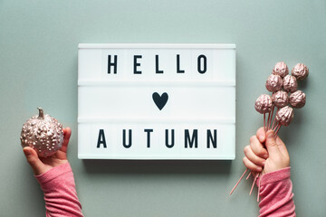 Lightbox with text Hello Autumn on silver grey paper background. Flat lay with female hands holding pink metallic pumpkin and small apples on sticks.