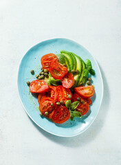 summer salad with ripe tomatoes and avocado with capers in a blue plate on the table
