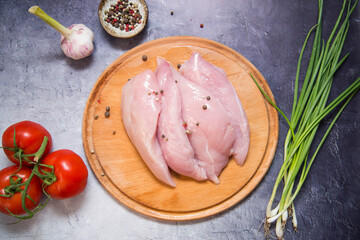Raw fillet of chicken for cooking some dish with pepper, garlic, green onion and tomatoes on the wooden plate. Top view. Grey background.