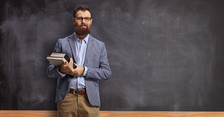 Bearded male teacher holding books and standing in front of a blackboard