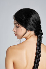 Cropped shot of a brunette lady with a wiglet made as a long black classic braid. The lady with naked shoulders is facing profile on the gray backdrop. 