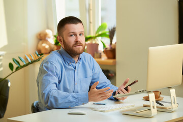 A man bewildered by his colleagues on an online business briefing in front of a laptop computer in his apartment. A guy in a blue shirt with a beard working remotely from home.