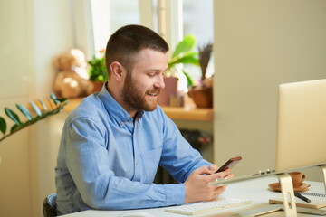 A happy young boss in a blue shirt searches the internet for news on a smartphone in front of a laptop computer in his apartment. A man with a beard working remotely from home. 