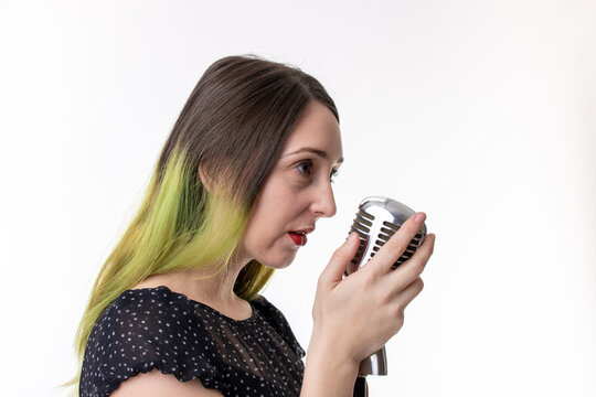 Studio photo of a young attractive woman with brown brunette hair and highlighted green hair, wearing a sexy black dress with red lip stick holding an old retro style microphone on a white background.