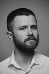 Fototapeta premium A close portrait of a bearded brutal man in a white shirt who is standing in front of a grey background. A serious guy is looking into the frame. A black and white photo. 