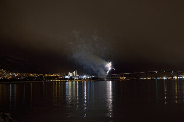 Naklejka na ściany i meble Beautiful firework on night sky in tromsoe city with bridge, cathedral and colorful reflection on the cold fjord water surface on new years eve