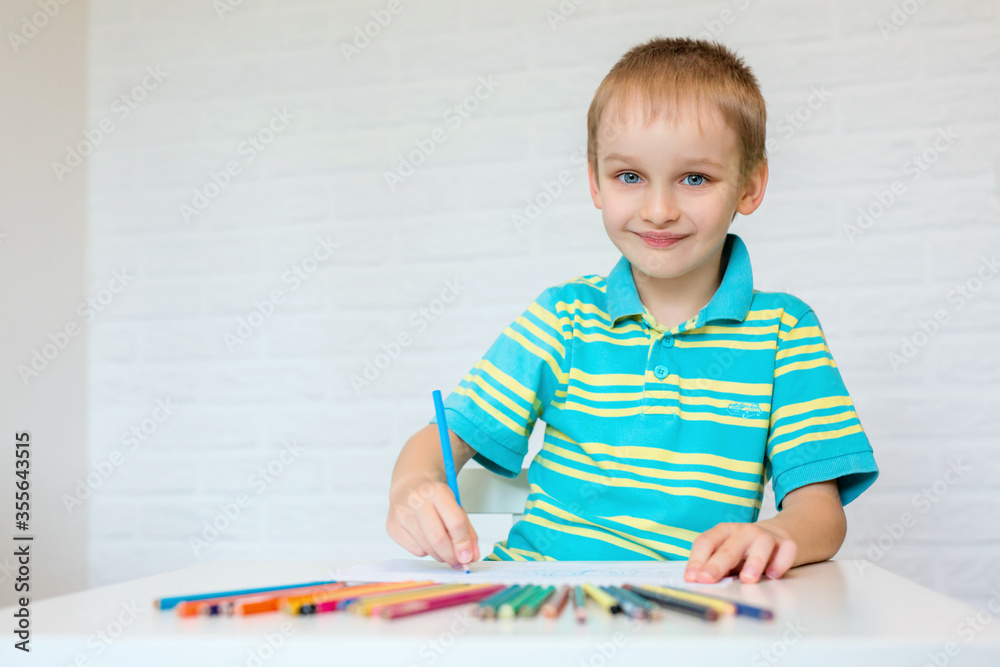 Wall mural a beautiful boy draws a cute drawing with colored pencils. a cute child is sitting at a table and ho