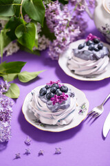 Obraz na płótnie Canvas Porcelain plate with meringue with whipped cream decorated with lilac flowers on violet background