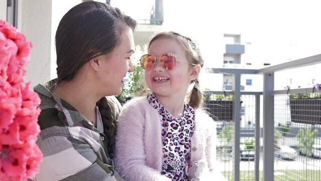 Young woman sits with her six-year-old daughter on the balcony. Mom and chaild are a lot of fun. Photographer is reflected in the mirror glasses of the girl.