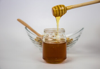 Glass jar with honey and a wooden stick on a white background. Pouring aromatic honey into jar, closeup.