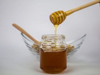 Glass jar with honey and a wooden stick on a white background. Pouring aromatic honey into jar, closeup.