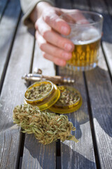 Table with marijuana grinder, marijuana bud, a smoking pipe and male hand holding a glass of beer...