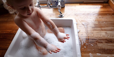 Funny small baby toddler playing in the sink Lockdown covid. Small baby playing in the bath sink. Bubbles and laughing. Adolescence. Baby taking a bath in the sink hygiene and skincare during lockdown