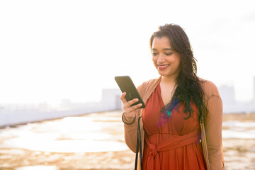 Happy young beautiful Indian woman using phone at rooftop of the building