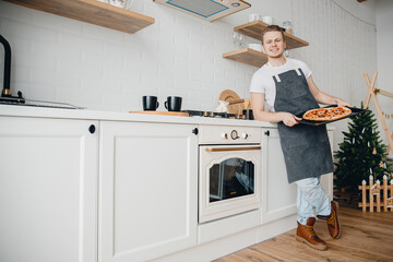 Homemade Italian pizza, handsome young male chef in dark apron holds baking sheet in bright...