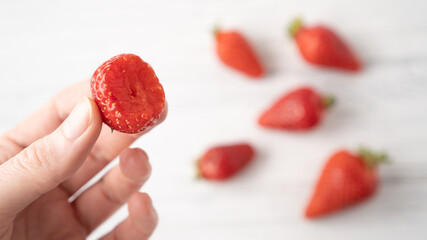 Fototapeta premium Hand holding bitten strawberry on white wooden background. Summer berry pleasure. Scattered strawberries