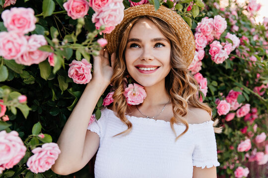 Winsome blue-eyed girl in summer hat posing in garden. Outdoor portrait of blithesome curly woman laughing with roses on background.