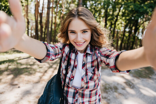 Inspired girl with light-brown hair making selfie in forest. Spectacular female model in checkered shirt expressing positive emotions.