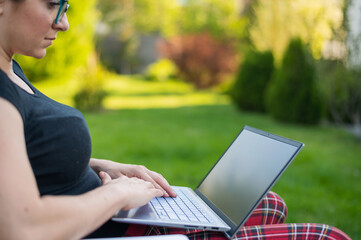 A red-haired woman sits in a park and types on the computer while shopping online. The girl maintains a social distance and works outdoors on a laptop. The student is studying remotely.