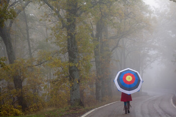 Mystical autumn foggy oak forest with empty road and a Girl with a red coat and an archery shooting...