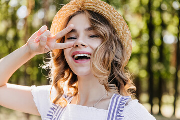 Close-up outdoor portrait of cheerful girl in straw hat. Enchanting young woman posing in forest with peace sign.