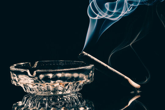 A cigar burning in glass ashtray with visible smoke on black background. Studio shot . Low key photo .