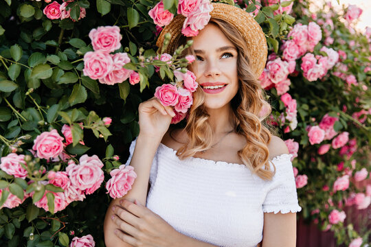 Blissful curly girl in white attire posing with smile near rose bush. Outdoor portrait of cheerful european lady in hat holding pink flower.