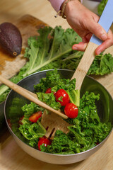 Close up of hands removing a kale and tomatoes salad in a metallic bowl with wooden spoons in a kitchen and an avocado in the background.