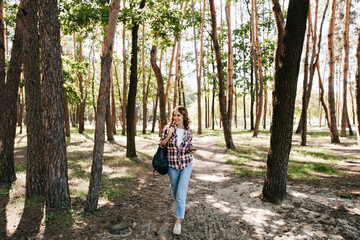 Full-length portrait of glad girl in jeans walking in forest. Winsome curly woman with backpack posing in park.