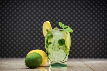 Classic lemonade in a skull-shaped glass at a marble board with pieces of ice on a spotted background. Halloween zombie cocktail soda, mint, lemon, ice and an unusual mug in the shape of a head