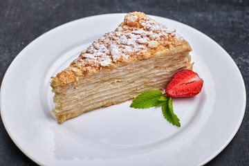 A piece of cake napoleon, on a white plate, with strawberries and mint, against a dark background