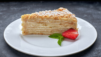 A piece of cake napoleon, on a white plate, with strawberries and mint, against a dark background