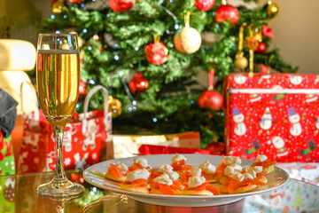 Fototapeta na wymiar Glass flute of champagne and smoked salmon canapes on a table with a Christmas tree and presents blurred in the background