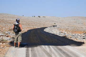 jeune homme en randonnée face à une route en bitume noir, regardant l'horizon et une vaste étendue désertique de pierres sur fond de ciel bleu.