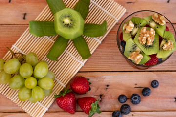 Above view of glass cup with healthy fresh and colorful fruit salad ready to eat. Wooden background with kiwi grapes strawberry and blueberries