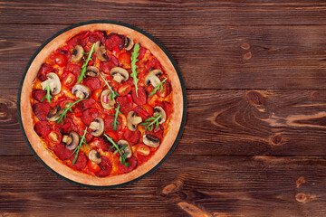 Delicious pizza hot with salami, arugula, cherry tomatoes, mushrooms and texas spice mix, on a round slate platter which is on wooden table, top view and copy space