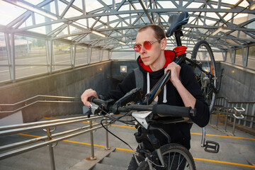 A young biker climbs the stairs of the underpass and carries a bicycle on his shoulder.