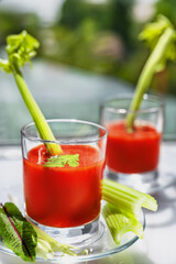 Two glasses of tomato juice with parsley and celery decorations, stand near the window, morning sunlight shines, shallow depth of field, selective focus. Natural drinks concept.