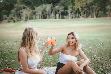 Happy friends on picnic in park, two young beautiful teen girls having fun and drinking orange juice while spending time together in the garden, woman outdoor friendship concept.